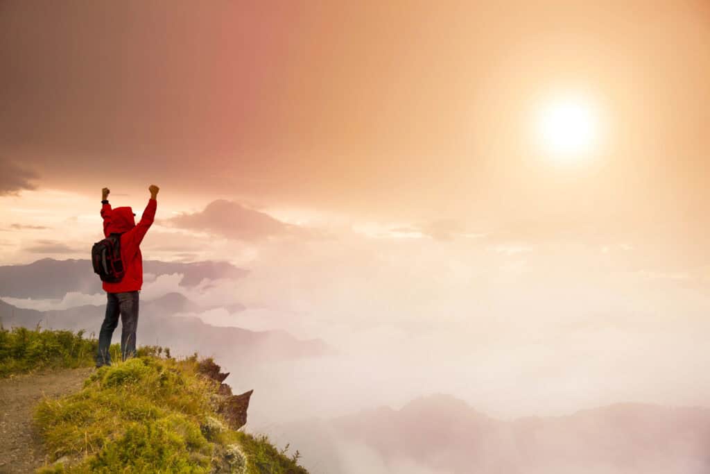 Ein Mann steht mit erhobenen Armen auf einem Berggipfel und blickt in die Ferne - Symbolbild für Resilienz im Job, Erfolg und das Erreichen von Zielen trotz Herausforderungen.