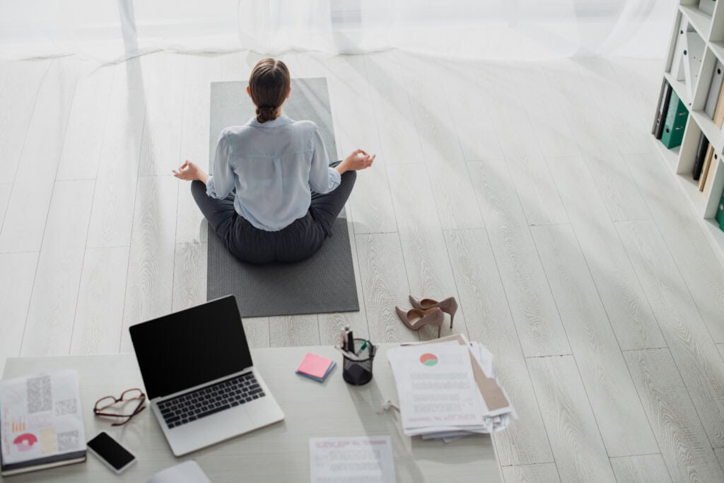 Eine Frau meditiert im Schneidersitz auf einer Matte in ihrem Büro. Dieses Bild symbolisiert, wie wichtig Meditation für Resilienz im Job und Stressabbau ist.