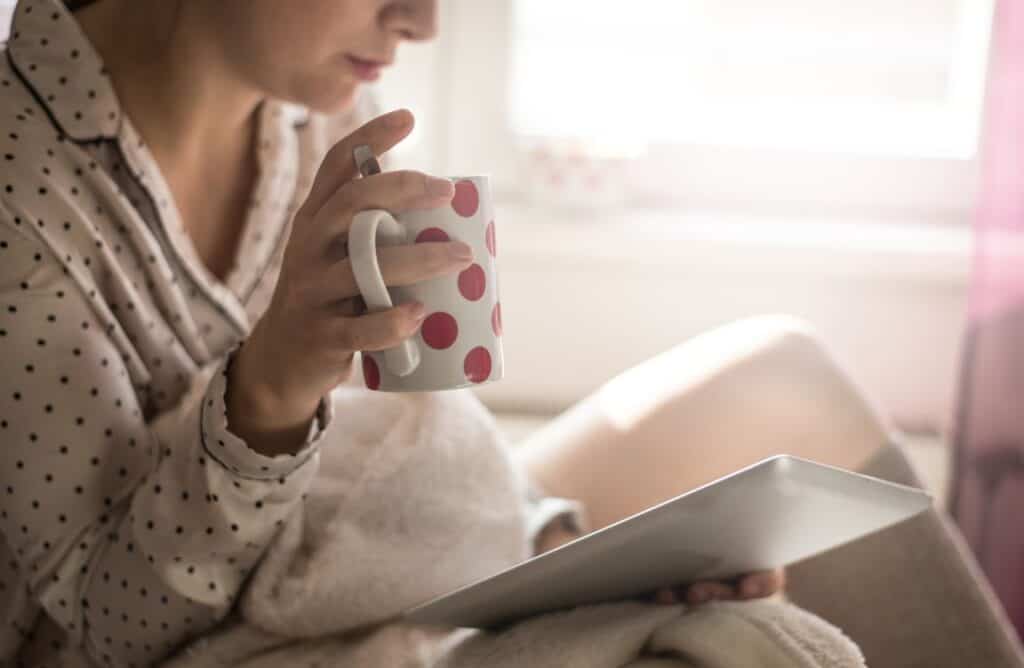 Frau mit einer Tasse Tee und einem Tablet, eingemummelt in eine Decke.