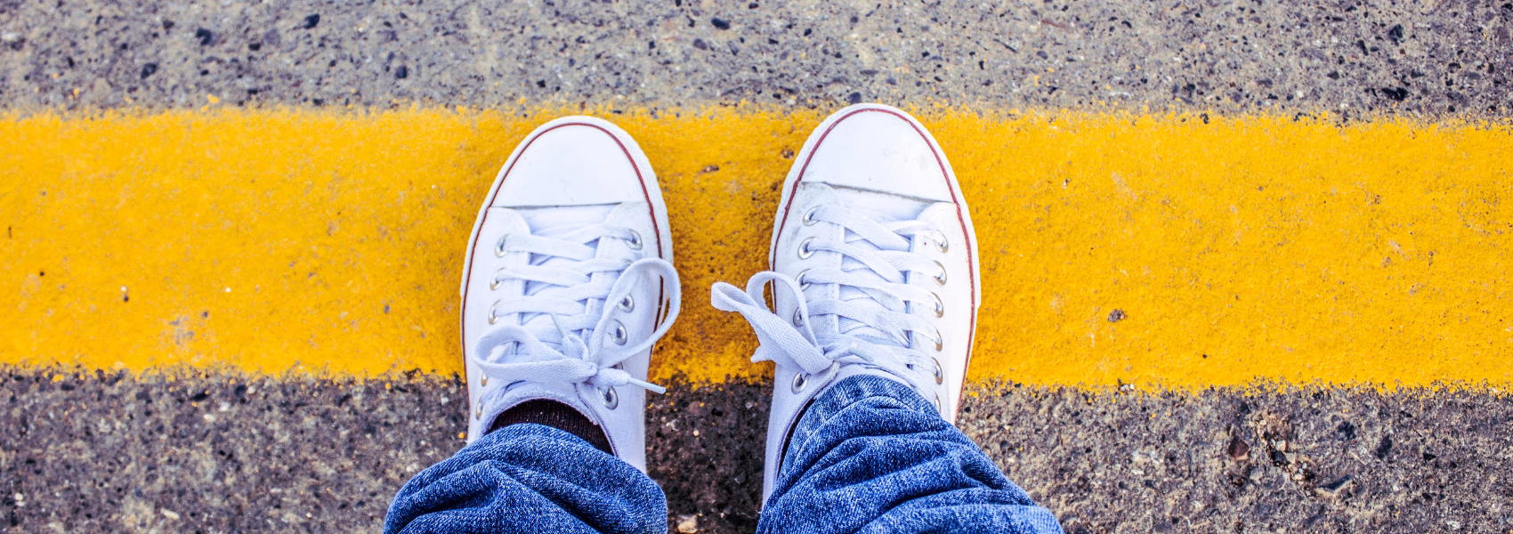 Von oben: Füße in weißen Sneakers und Jeans stehen hinter einer dicken gelben Linie auf Asphalt – klares Symbol für Grenzen setzen nach Trennung.