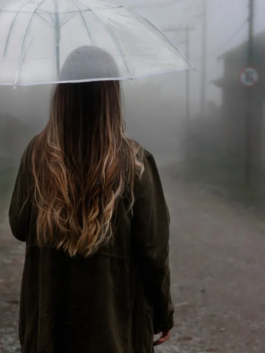 Frau von hinten mit Regenschirm auf einer Straße im Nebel, Symbol für Orientierungslosigkeit.